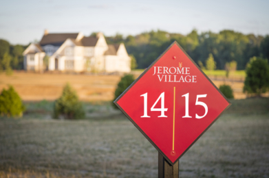 Jerome Village lot number sign red with white numbers 14 and 15 with a view of an empty home lot, treeline, and large custom home with a vast amount of trees behind the large home lot line.